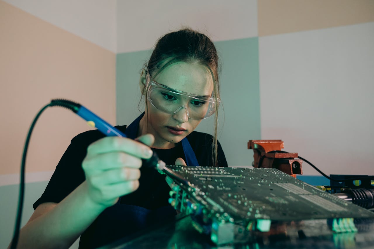 Young woman wearing safety glasses soldering a circuit board in a workshop setting.